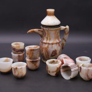 Pode incluir: Um conjunto de café em ônix, incluindo uma cafeteira alta com tampa e várias xícaras pequenas. As peças são em tons de marrom, branco e bege, com uma aparência marmorizada. O conjunto é exibido em uma superfície escura.