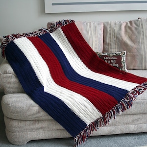May include: A handmade crochet blanket in a patriotic color scheme of red, white, and blue. The blanket features vertical stripes and a fringed edge. It is draped over a neutral-toned sofa.