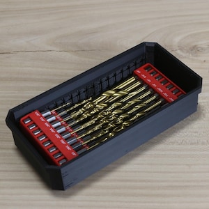 May include: A black plastic storage box with a red and white label containing a set of gold drill bits.