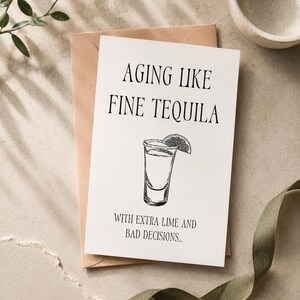 Può includere: Un biglietto d'auguri bianco con testo nero che recita AGING LIKE FINE TEQUILA WITH EXTRA LIME AND BAD DECISIONS. Sotto il testo è presente un disegno a linee in bianco e nero di un bicchiere da shot di tequila con una fetta di lime.