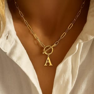 May include: Gold-coloured initial necklace with an "A" pendant. The necklace has a toggle clasp and a chain with elongated links. The jewellery is worn over a white collared shirt.
