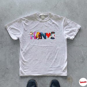 May include: White t-shirt with the word "KANYE" in colorful, stylized letters. The letters feature various cartoon images and designs. The shirt has a crew neck and short sleeves. The shirt is on a gray concrete surface.