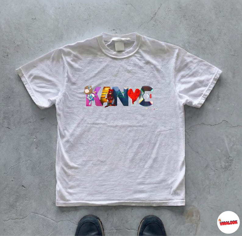 May include: White t-shirt with the word "KANYE" in colorful, stylized letters. The letters feature various cartoon images and designs. The shirt has a crew neck and short sleeves. The shirt is on a gray concrete surface.