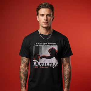 Peut inclure: Un t-shirt noir à manches courtes avec une impression graphique portant le texte 'I am the Third Revelation! I am who the Lord has chosen!' au-dessus d'une scène de deux personnages, avec le mot 'Drainage' en grande police gothique blanche.