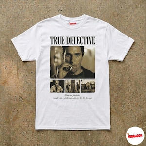 Op de afbeelding: Wit T-shirt met de tekst "TRUE DETECTIVE" boven een sepia-afbeelding van een rokende man. Onder de afbeelding staan drie kleinere afbeeldingen en de tekst "Time is a flat circle."