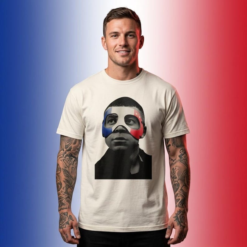 K&ouml;nnte beinhalten: Ein cremefarbenes Kurzarm-T-Shirt mit einem Grafikdruck einer Person, die eine Maske mit den blauen, wei&szlig;en und roten Streifen der franz&ouml;sischen Flagge tr&auml;gt. Die Person auf der Grafik hat einen dunklen Teint und tr&auml;gt ein dunkles Oberteil.