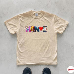May include: A cream-colored t-shirt with the word "KANYE" in colorful, stylized letters. The letters feature various designs, including a heart and cartoon figures. The shirt is made of a soft material and has a classic crew neck.