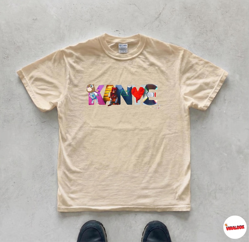May include: A cream-colored t-shirt with the word "KANYE" in colorful, stylized letters. The letters feature various designs, including a heart and cartoon figures. The shirt is made of a soft material and has a classic crew neck.