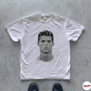 Può includere: T-shirt bianco con un ritratto in bianco e nero di un uomo e una firma. La maglietta è appoggiata su una superficie grigia. Ha maniche corte e girocollo. L'immagine è in bianco e nero.