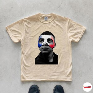 K&ouml;nnte beinhalten: Ein beiges Kurzarm-T-Shirt mit einem schwarz-wei&szlig;en Fotodruck einer Person, die eine Maske in den Farben Blau, Wei&szlig; und Rot der franz&ouml;sischen Flagge tr&auml;gt. Das T-Shirt hat einen Rundhalsausschnitt und ein klassisches Design.
