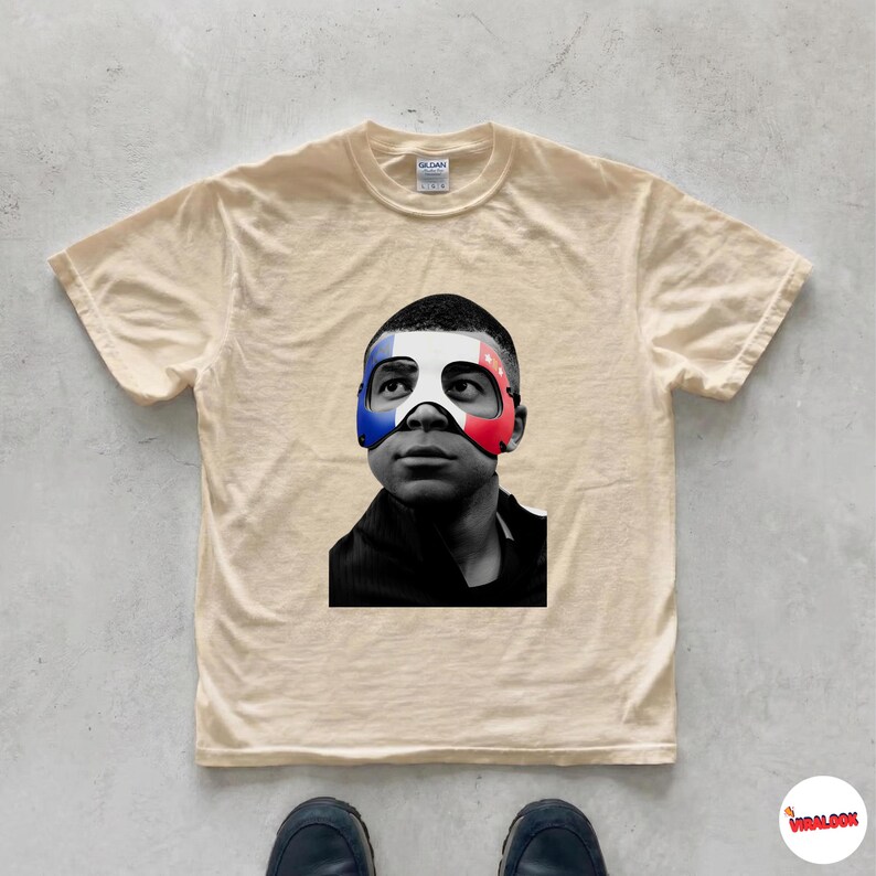 K&ouml;nnte beinhalten: Ein beiges Kurzarm-T-Shirt mit einem schwarz-wei&szlig;en Fotodruck einer Person, die eine Maske in den Farben Blau, Wei&szlig; und Rot der franz&ouml;sischen Flagge tr&auml;gt. Das T-Shirt hat einen Rundhalsausschnitt und ein klassisches Design.