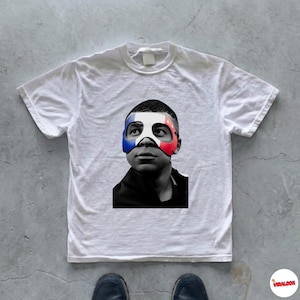 K&ouml;nnte beinhalten: Ein wei&szlig;es Kurzarm-T-Shirt mit einem schwarz-wei&szlig;en Fotodruck einer Person, die eine Maske mit den blauen, wei&szlig;en und roten Streifen der franz&ouml;sischen Flagge &uuml;ber Augen und Nase tr&auml;gt, vor einem schlichten wei&szlig;en Hintergrund.