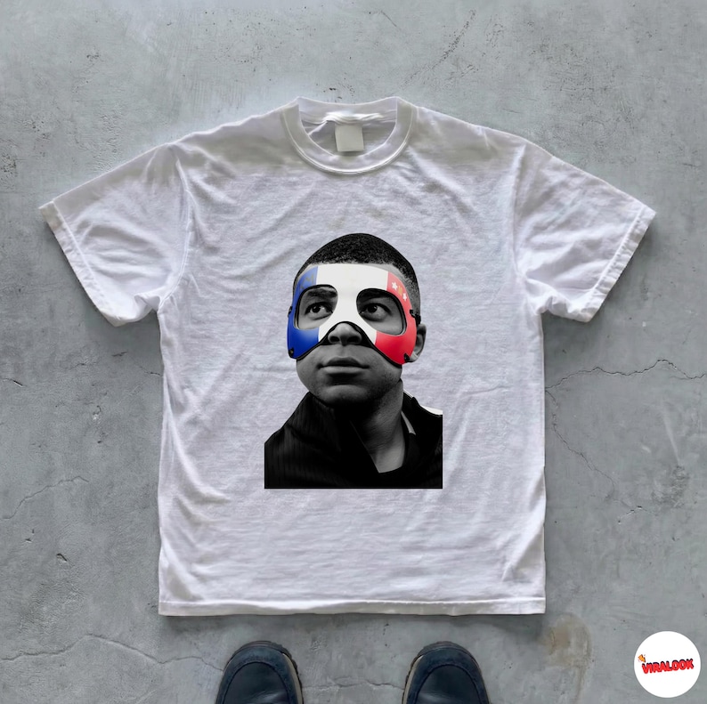 K&ouml;nnte beinhalten: Ein wei&szlig;es Kurzarm-T-Shirt mit einem schwarz-wei&szlig;en Fotodruck einer Person, die eine Maske mit den blauen, wei&szlig;en und roten Streifen der franz&ouml;sischen Flagge &uuml;ber Augen und Nase tr&auml;gt, vor einem schlichten wei&szlig;en Hintergrund.