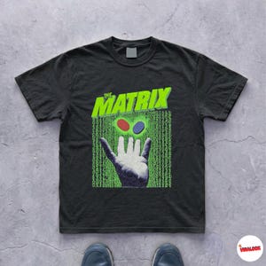 Peut inclure: Un t-shirt noir à manches courtes avec une impression graphique représentant une main tendant vers une pilule rouge et une pilule bleue. Au-dessus de la main, le texte THE MATRIX en lettres vert vif sur un fond de code numérique vert.
