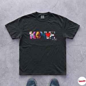 May include: Black t-shirt with the name "KANYE" in colorful block letters. The letter "Y" is a red heart. The word "west" is written below the name in a smaller font. The shirt is made of a soft material.