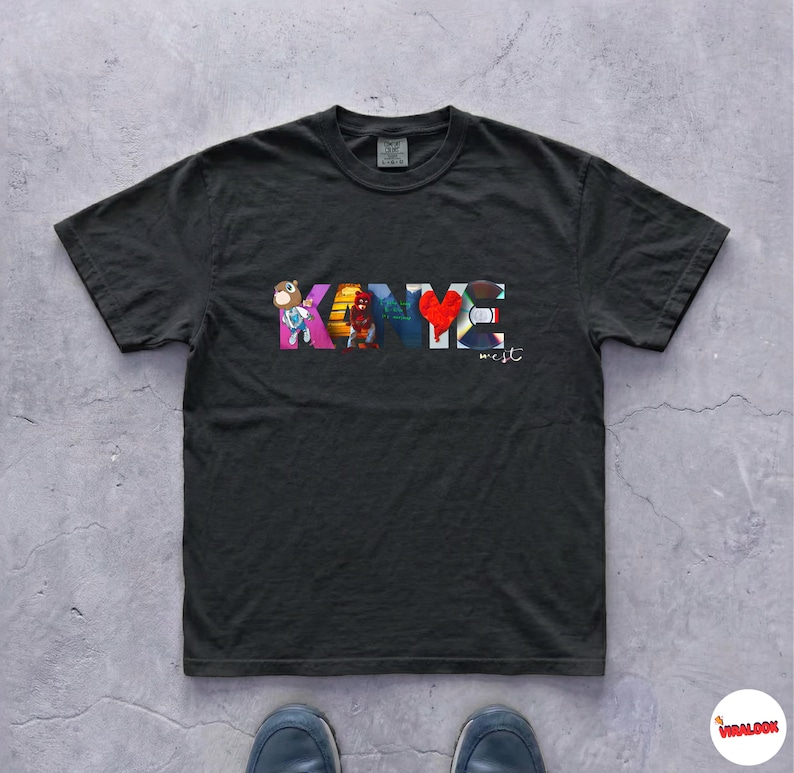 May include: Black t-shirt with the name "KANYE" in colorful block letters. The letter "Y" is a red heart. The word "west" is written below the name in a smaller font. The shirt is made of a soft material.