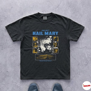 Peut inclure: T-shirt noir avec l'inscription "PROJECT HAIL MARY" en bleu. Le motif comprend une image centrale et plusieurs petites images dans des carrés jaunes. Le texte "Amazed! Amazed! Amazed!" est en bas du motif.