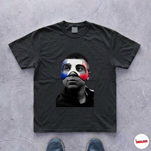 K&ouml;nnte beinhalten: Ein schwarzes Kurzarm-T-Shirt mit einer mittigen Grafik eines Gesichts in Schwarz-Wei&szlig;. Die Person tr&auml;gt eine Maske mit blauen, wei&szlig;en und roten Streifen &uuml;ber dem Augen- und Nasenbereich. Das T-Shirt hat einen klassischen Rundhalsausschnitt.