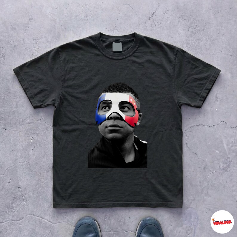 K&ouml;nnte beinhalten: Ein schwarzes Kurzarm-T-Shirt mit einer mittigen Grafik eines Gesichts in Schwarz-Wei&szlig;. Die Person tr&auml;gt eine Maske mit blauen, wei&szlig;en und roten Streifen &uuml;ber dem Augen- und Nasenbereich. Das T-Shirt hat einen klassischen Rundhalsausschnitt.