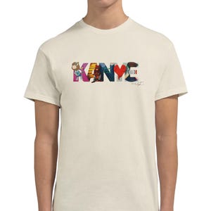 May include: Cream-colored t-shirt featuring the word "KANYE" in a colorful, artistic design. The letters are filled with various images and patterns, including a bear, a figure, and a heart. A signature is visible below the word.