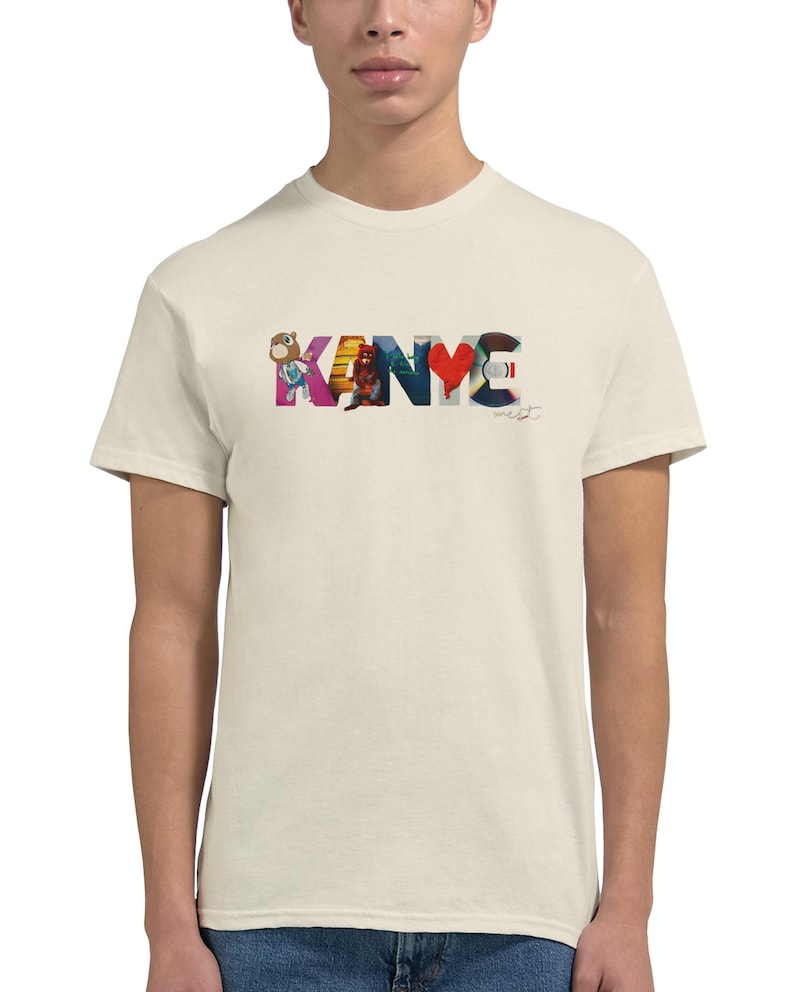 May include: Cream-colored t-shirt featuring the word "KANYE" in a colorful, artistic design. The letters are filled with various images and patterns, including a bear, a figure, and a heart. A signature is visible below the word.
