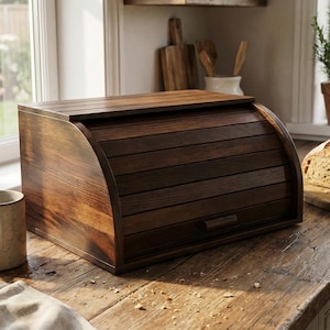 Könnte beinhalten: Eine dunkelbraune Brotdose aus Holz mit Rollladen-Deckel steht auf einer rustikalen Holz-Arbeitsplatte. Die Dose hat eine gebogene Front und einen kleinen Griff. Ein Brotlaib liegt auf einem Holzschneidebrett. Die Küche umfasst ein Fenster und eine Tasse.