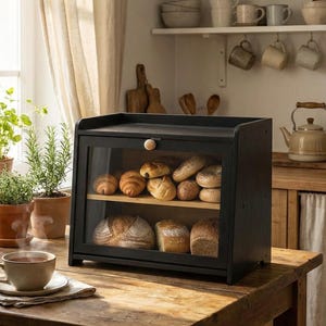 Puede incluir: Una panera de madera negra con puerta de cristal, que muestra varios tipos de pan. La panera está sobre una mesa de madera, junto a una ventana con plantas y una taza de té. El ambiente de la cocina es rústico.