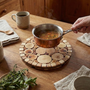 May include: A copper saucepan filled with soup sits on a wooden trivet made of wood slices. The trivet is on a wooden table, with a mug, spoon, and sprigs of herbs nearby. The kitchen setting has wooden cabinets.