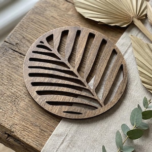 Peut inclure: Un dessous de verre rond en bois avec un motif de feuille découpée. Le dessous de verre est en bois brun foncé et repose sur une table en bois. Des feuilles de palmier séchées et de la verdure sont en arrière-plan.