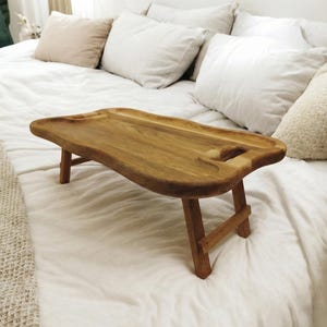 May include: Wooden bed tray with a natural finish. The tray has an organic shape with a handle cutout. Supported by folding legs, it's ideal for breakfast in bed or working. The tray's dimensions are approximately 60 cm long.