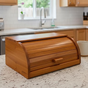 May include: A wooden bread bin with a roll-top lid and a small handle. The bin is made of light brown wood and sits on a speckled countertop in a kitchen. A mug and a fruit basket are nearby.