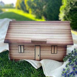 May include: A wooden miniature house with a light brown roof and walls. The house has two windows, a door, and a small rectangular piece above the door. The house is sitting on a white cloth and green grass.