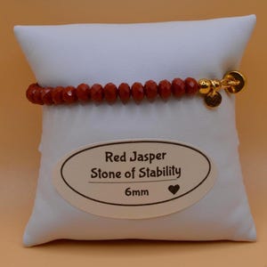 May include: Red Jasper beaded bracelet with gold-colored charms. The bracelet is displayed on a white pillow with a label that reads "Red Jasper Stone of Stability 6mm". The red beads are approximately 0.24 inches in diameter.
