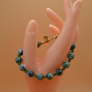 May include: A beaded bracelet featuring large, round, green and white speckled stones interspersed with smaller green beads. The bracelet is held by a hand and has a gold clasp. The bracelet is a piece of jewelry.