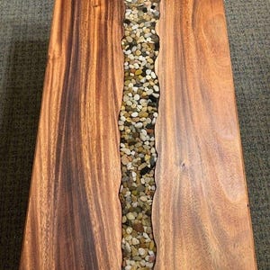 May include: A rectangular wooden coffee table with a river of small, multicolored stones running down the centre. The wood has a rich, reddish-brown colour and visible grain. The stone river adds a natural, textured contrast.