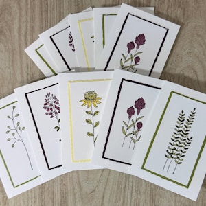 Puede incluir: Un conjunto de tarjetas de felicitación blancas con ilustraciones botánicas. Cada tarjeta presenta un diseño floral diferente, incluyendo plantas moradas, amarillas y verdes, enmarcadas por bordes de colores. Las tarjetas están dispuestas sobre una superficie de madera clara.