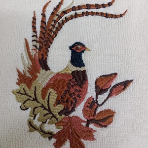 May include: Embroidered pheasant design in shades of brown, orange, and green on a white background. The bird is surrounded by leaves, with detailed feathering and a black head. The image is a close-up of the embroidery.