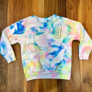 Puede incluir: Una sudadera blanca con un patrón tie-dye en tonos de azul, rosa, amarillo y verde. La sudadera tiene una letra "J" de purpurina dorada en la parte delantera.
