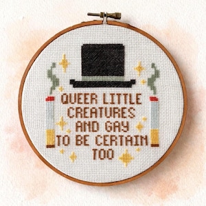 May include: A cross-stitch embroidery in a round wooden hoop. The design features a black top hat, two stylized cigarettes, and the text "QUEER LITTLE CREATURES AND GAY TO BE CERTAIN TOO" in brown letters, with yellow star accents.