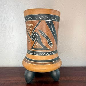 May include: A cylindrical, handmade ceramic vase with a brown and tan color scheme. The vase features black geometric designs and rests on three black feet. The design includes triangles, lines, and wave patterns.