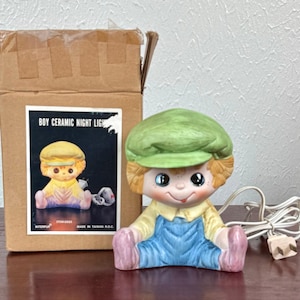 May include: A ceramic night light shaped like a seated boy wearing a green cap and blue overalls. The night light has a smiling face with rosy cheeks and is next to its original box, which reads "BOY CERAMIC NIGHT LIGHT."