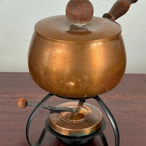 May include: A vintage copper fondue pot with a wooden handle and lid knob. The pot sits on a black metal stand with a burner. The copper has a warm, aged patina, suggesting it's a well-loved kitchen item.