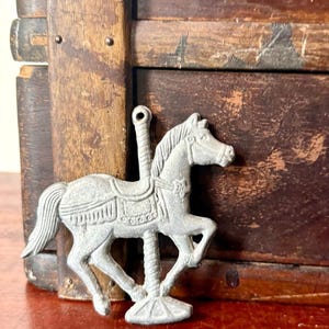 May include: A flat, silver-toned metal carousel horse ornament featuring a textured mane, a saddle with decorative fringe, and a star detail on the bridle. The horse is attached to a central pole with a circular base and a loop at the top for hanging.