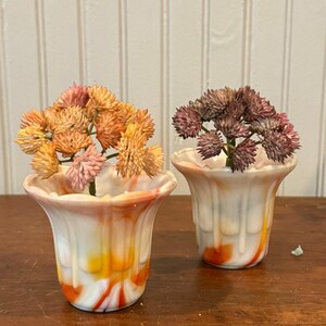 May include: Two small, fluted glass vases with a white base and swirling orange and red patterns. Each vase contains a small artificial plant with clustered, textured flower heads in shades of peach and muted purple, respectively.