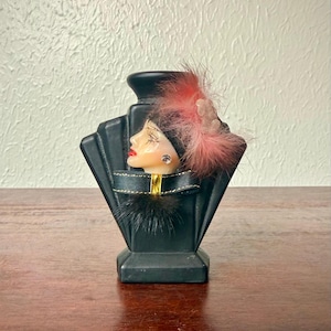 May include: Black art deco perfume bottle with a woman's face. The bottle has a fan-shaped design and is adorned with a pink feather and a gold ribbon. The woman's face has red lips and a small earring.