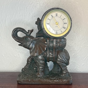 May include: A bronze-colored elephant figurine with a clock on its back. The clock has a gold-colored bezel and a cream-colored face with black Roman numerals. The elephant is detailed with wrinkles and a trunk raised in the air.