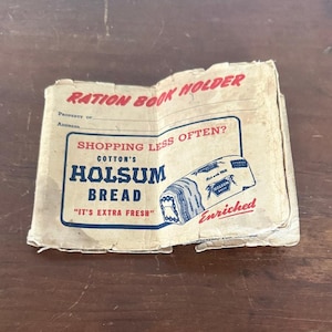 May include: A vintage, off-white paper ration book holder featuring red and blue text. The cover reads RATION BOOK HOLDER, SHOPPING LESS OFTEN?, COTTON'S HOLSUM BREAD, IT'S EXTRA FRESH, and ENRICHED, with a graphic illustration of a loaf of bread.