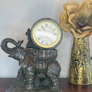 May include: A bronze-colored elephant figurine with a clock face on its back. The clock has Roman numerals and a gold-colored bezel. The elephant is standing on a textured base, and a decorative vase with a flower is to the right.