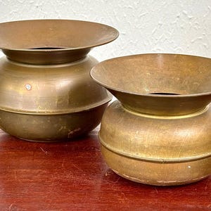 May include: Two antique brass planters with a wide, flared rim and a rounded body. The planters have a warm, golden-brown color and sit on a wooden surface. The design is simple and elegant, suitable for various plants or decorative purposes.