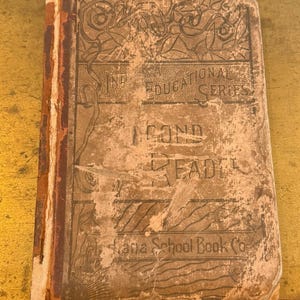 May include: An antique book with a worn, tan cover. The cover features the text "IND EDUCATIONAL SERIES" and "2ND READER". The book's title is partially obscured by wear and tear. The book is from the "Indiana School Book Co."